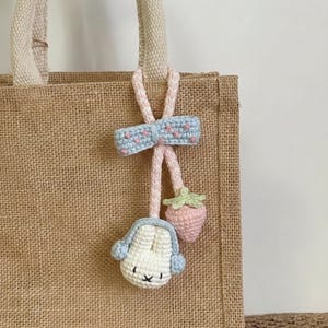 Puede incluir: Un charm de bolso de ganchillo hecho a mano con un conejito blanco con orejeras azules, una fresa rosa y un lazo azul con detalles rosas. El charm está sujeto a un bolso tote de arpillera con un asa beige.