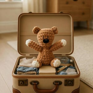 Può includere: Una valigia beige aperta, con finiture in pelle marrone, rivela un orsacchiotto marrone all'uncinetto con zampe bianche. La valigia è piena di vestiti piegati, un libro e oggetti da viaggio.