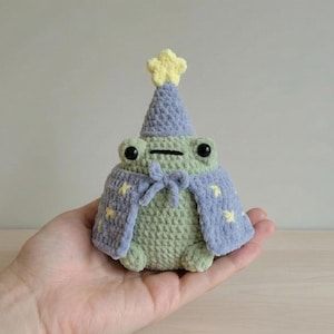 Puede incluir: Figura de rana de ganchillo con sombrero y capa de mago. La rana es verde con ojos negros y una boca pequeña cosida. El sombrero y la capa son de color azul claro con detalles de estrellas amarillas. Una estrella amarilla adorna la parte superior del sombrero.