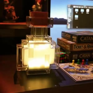 Könnte beinhalten: Eine klare, pixelartige Lampe in Form einer Trankflasche mit warmem, gelbem Licht. Die Lampe hat einen roten Deckel und steht auf einer dunklen Oberfläche. Im Hintergrund sind Brettspiele zu sehen, im Stil eines Videospiels.