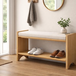 May include: A light wood entryway bench with a cream cushion and a lower shelf. Two pairs of shoes are on the shelf. A small vase with greenery sits on the bench. A woven basket and a door are in the background.