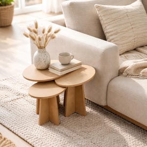 Puede incluir: Un salón de tonos neutros con un sofá beige, una alfombra y un conjunto de tres mesas auxiliares de madera. Un jarrón con flores secas, una taza y libros están sobre las mesas.