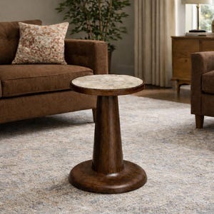 May include: A dark brown, wooden side table with a round top and a pedestal base. The tabletop has a light-colored, speckled surface. The table is placed on a patterned rug in a living room setting.