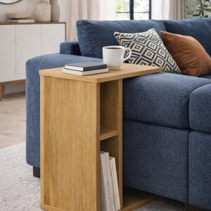 May include: A wooden side table with a natural finish, featuring a tabletop and a lower shelf for storage. A white coffee cup and a book rest on the table. The table is positioned next to a blue sofa, with a rug beneath.