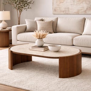 May include: A modern living room scene featuring a beige sofa with decorative pillows, a round wooden side table with a lamp, and an oval coffee table. The coffee table has a light-colored top and a dark wood base. A cream-colored rug covers the floor.