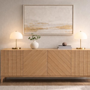 May include: A light wood sideboard with a geometric design, topped with two lamps, a white vase with greenery, and books. A framed abstract painting hangs above the sideboard. A potted plant is on the left.