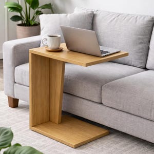 May include: A light brown, C-shaped side table with a laptop and a cup of coffee on top. The table is made of wood and sits next to a grey sofa. The laptop is open, and the coffee cup is white.
