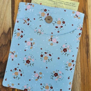 May include: A light blue fabric book sleeve with a floral pattern in pink, brown, and peach. The sleeve has a pocket and a button closure. A yellow card with text is partially visible inside the sleeve.