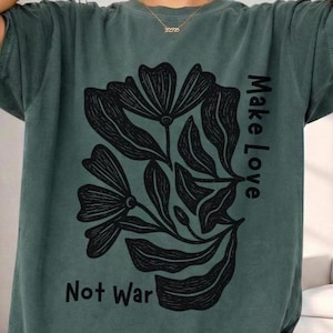 Może przedstawiać: Ciemnozielona koszulka z czarnym kwiatowym wzorem i napisami "Make Love" i "Not War". Koszulka ma krótkie rękawy i okrągły dekolt.