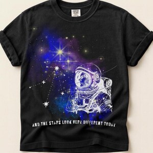 May include: Black t-shirt featuring a white illustration of an astronaut in a spacesuit, set against a vibrant nebula of blues, purples, and yellows. The text "AND THE STARS LOOK VERY DIFFERENT TODAY" is printed below the graphic.