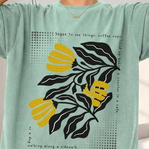 Puede incluir: Camiseta verde claro con un diseño floral. El diseño presenta flores amarillas, hojas negras y el texto: "I began to see things: coffee cups lined up behind a counter in a cafe. or a dog walking along a sidewalk."