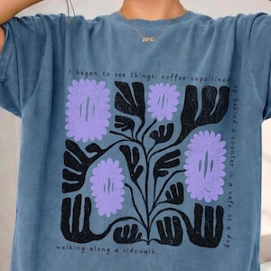 Puede incluir: Camiseta azul con un diseño floral en negro y lavanda. La camiseta presenta el texto "I began to see things: coffee cups lined up behind a counter in a cafe. or a dog walking along a sidewalk."