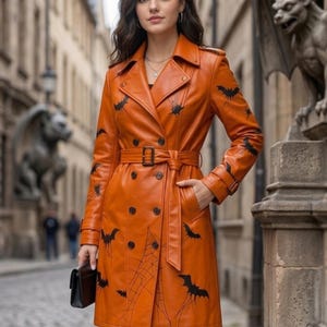 Könnte beinhalten: Ein orangefarbener, zweireihiger Trenchcoat mit einem schwarzen Fledermaus- und Spinnennetz-Druck. Der Mantel verfügt über einen passenden Bindegürtel mit Schnalle, fallendes Revers, lange Ärmel mit Riegeln an den Manschetten und zwei Taschen.
