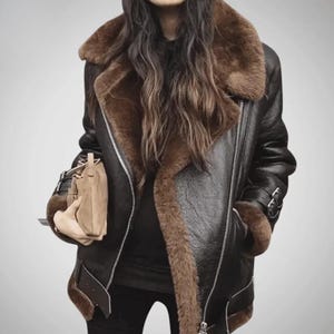 Puede incluir: Chaqueta de cuero negra con forro y cuello de borrego marrón. La chaqueta tiene cierre de cremallera, puños con hebillas y un ribete de borrego marrón. Un bolso de mano beige se sostiene en la mano izquierda.