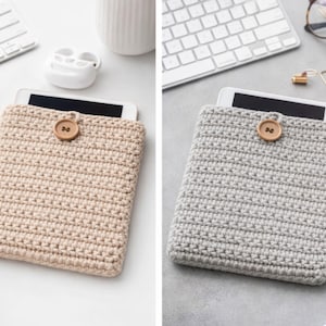May include: Two crochet tablet cases, one in beige and one in grey, each secured with a wooden button. The cases are handmade, likely from thick yarn. Tablets are partially visible within the cases.