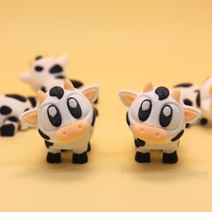 Puede incluir: Varias pequeñas figuras de vaca blancas con manchas negras y rasgos de dibujos animados. Las vacas tienen ojos grandes y negros, narices color melocotón y pequeñas pezuñas negras. El fondo es de color amarillo claro.