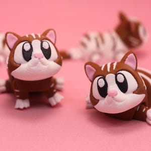 May include: Three small, brown and white cat figurines with large black eyes and pink noses. Each cat has a rounded head, pointed ears with pink inner details, and white markings on the face, paws, and back, featuring a smooth, layered texture.
