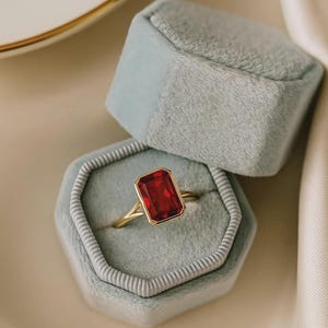 May include: A gold ring featuring a large, rectangular, emerald-cut red gemstone set in a bezel setting. The ring is displayed inside an open, hexagonal, light blue velvet jewelry box with a soft, plush interior lining and a matching lid.