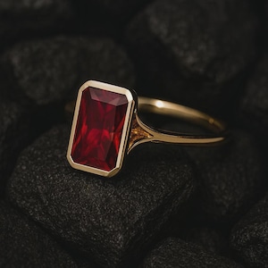May include: A gold ring with a rectangular, deep red gemstone. The gemstone is set in a gold bezel, and the ring's band has a simple, elegant design. The ring is shown against a dark, textured background.