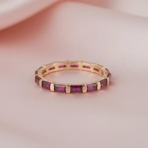 Op de afbeelding: Een goudkleurige ring met een doorlopende band van rechthoekige, dieproze edelstenen. De ring is geplaatst tegen een zachte, roze achtergrond, waardoor het contrast tussen het metaal en de stenen wordt benadrukt. Het ontwerp is elegant en modern.