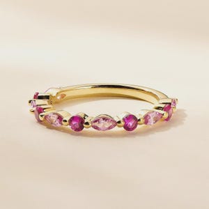 Puede incluir: Un anillo de oro con gemas rosas redondas y en forma de marquesa alternadas. El anillo tiene una banda delicada y está engastado con una serie de piedras rosas vibrantes. Las gemas son una mezcla de tonos rosas claros y oscuros.