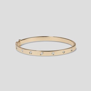 Puede incluir: Una pulsera de brazalete dorada con varias piedras preciosas pequeñas y transparentes engastadas en la banda. La pulsera tiene un cierre de bisagra y un acabado liso y pulido. Las piedras preciosas están espaciadas uniformemente.