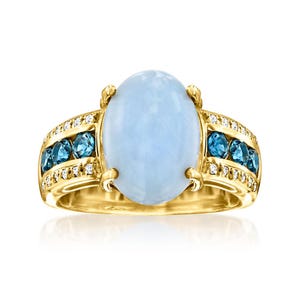 May include: A gold ring featuring a large, oval, cabochon-cut light blue gemstone at the center. The band is accented with rows of smaller, round, deep blue gemstones and tiny clear stones set along the sides of the central setting.