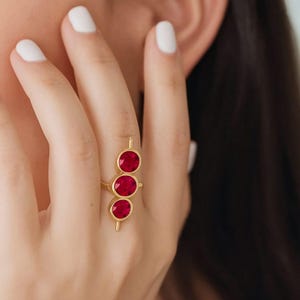 May include: A gold ring with three bright red gemstones. The ring has a vertical design, with the gemstones set in individual bezels. The ring is displayed on a finger, showcasing the jewellery's elegant simplicity. The gemstones are a vibrant red.