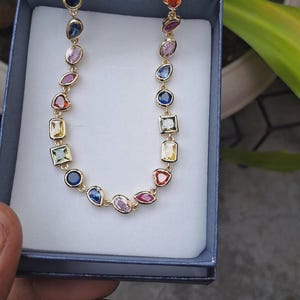 Könnte beinhalten: Eine goldfarbene Halskette mit einer Reihe farbiger Edelsteine in verschiedenen Formen, darunter rund, oval und rechteckig. Die Edelsteine sind in einzelnen Goldfassungen gefasst und in einem linearen Muster angeordnet. Die Halskette wird in einer blauen Schmuckschatulle präsentiert.