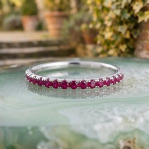 Può includere: Un sottile anello a fascia in argento con una fila continua di piccoli rubini rotondi di un vivido colore rosso rosato, incastonati in una montatura a griffe condivisa. Il metallo ha una finitura lucida e riflettente, con pietre uniformi.