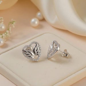 May include: A pair of silver stud earrings. Each earring has a heart-shaped design with a wing motif, edged with small, clear gemstones. The earrings are presented on a cream-coloured surface.