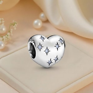 May include: A silver heart-shaped charm with star-shaped cutouts and small blue gemstones. The charm has the word "DISNEY" engraved on the side. The charm is displayed on a light beige surface.
