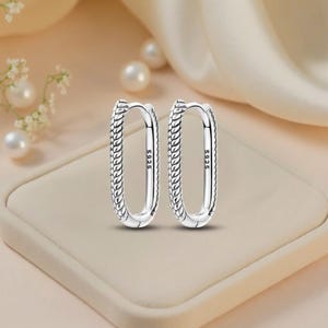 May include: A pair of silver oval hoop earrings. The earrings have a textured, braided design on the outer edge and are marked with "S925". The earrings are displayed on a beige surface with pearls and white flowers in the background.