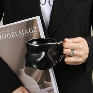 May include: A black ceramic mug with a unique, textured design and a silver-colored rim and handle. The mug is held over a magazine with the text "MODELMAGZ". The person is wearing a black blazer.