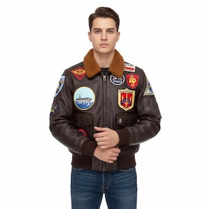 May include: A brown leather bomber jacket with various patches, a brown fur collar, and ribbed cuffs and hem. The jacket features a zip closure and two front pockets. Patches include text and emblems. The model is wearing blue jeans.
