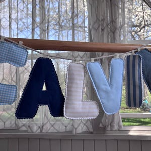 Puede incluir: Una pancarta con la palabra "CALVIN" cuelga frente a una ventana. Las letras son de tela en varios tonos de azul, con patrones como cuadros y rayas. La pancarta está sujeta a una barra de madera.