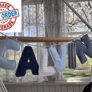 Op de afbeelding: Een stoffen banner met de naam "CALVIN", waarbij elke letter een ander patroon en kleur heeft, waaronder blauw ruitjes, marineblauw en strepen. De banner hangt aan een houten stok. Een stempel "RUSH ORDER UPGRADE" is zichtbaar.
