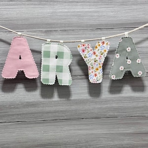 Op de afbeelding: Een decoratieve banner met de tekst "ARYA" met viltletters. De letters zijn in verschillende patronen: roze, groen geruit, bloemen en saliegroen met witte madeliefjesaccenten. De banner is geregen aan een wit touw tegen een grijze houten achtergrond.