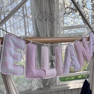 May include: A fabric banner spelling the name ELLIANA in individual stuffed letters. Each letter features a different shade of pink, including gingham, solid, floral, and paisley patterns, all suspended from a horizontal wooden rod by rope.