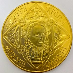 Può includere: Una moneta color oro con l'iscrizione "ELIZABETH REGINA" e numeri romani. La moneta presenta un ritratto di donna con colletto arricciato e corona all'interno di un disegno a forma di diamante.
