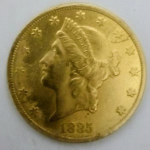 May include: A gold coin with a profile of a woman wearing a crown, dated 1885. The coin's design includes stars and floral details around the rim. The coin has a bright, reflective surface, typical of gold coinage.