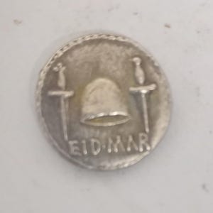 Puede incluir: Una moneda plateada con un diseño detallado. La moneda presenta un yelmo entre dos espadas y la inscripción "EID MAR". La moneda es circular con un borde texturizado, sobre un fondo blanco.