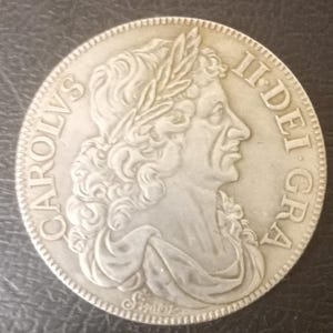 May include: A silver coin featuring a profile of a man wearing a laurel wreath. The coin has the inscription "CAROLVS II DEI GRA" around the edge. The coin has a detailed design and a slightly worn appearance.