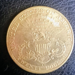 Könnte beinhalten: Eine goldene Münze mit aufwendigen Details. Die Worte "UNITED STATES OF AMERICA" sind am oberen Rand eingraviert. Die Münze zeigt einen Adler mit einem Schild, die Worte "IN GOD WE TRUST" und die Bezeichnung "TWENTY DOLLARS".