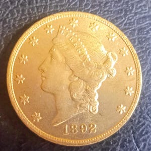 Może przedstawiać: Złota moneta z 1892 roku, przedstawiająca profil kobiety ze słowem "LIBERTY" nad głową. Moneta ma szczegółową krawędź i jest otoczona małymi wzorami w kształcie gwiazd.