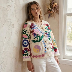 Może przedstawiać: Kremowa haftowana kurtka z kolorowymi wzorami kwiatowymi, owocowymi i symbolami. Kurtka posiada zapinany na guziki przód, dwie kieszenie i symbol Yin Yang. Haft zawiera różowe, niebieskie, czerwone i zielone detale.