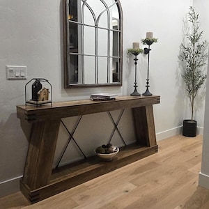 May include: A wooden console table with a decorative arched mirror above it. The table has a lower shelf and metal cross supports. Two black candle holders and a small plant are on the table. A sign on the wall reads "ALWAYS PRAY TO HAVE".