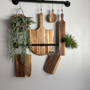 May include: A kitchen wall display featuring a black metal rod with hanging wooden cutting boards of various shapes and sizes. A small hanging basket with greenery and a small plant are also suspended from the rod. The cutting boards are made of light brown wood.