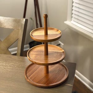 Puede incluir: Una bandeja de servicio de madera de tres niveles con un poste central vertical y tres plataformas circulares. La madera tiene un tono marrón medio cálido. La bandeja está diseñada para exhibir o servir alimentos.
