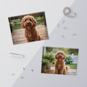 May include: Two spiral-bound 2026 calendars featuring a Cockapoo dog. One calendar shows the dog on a wooden deck, the other on a porch with potted plants. The calendars are on a white surface with office supplies.